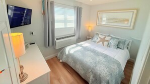 A bedroom with a bed, a TV, a desk, a lamp, a window with curtains, and a wall-mounted picture. - The Breakaway Beach Condo (North Wildwood)
