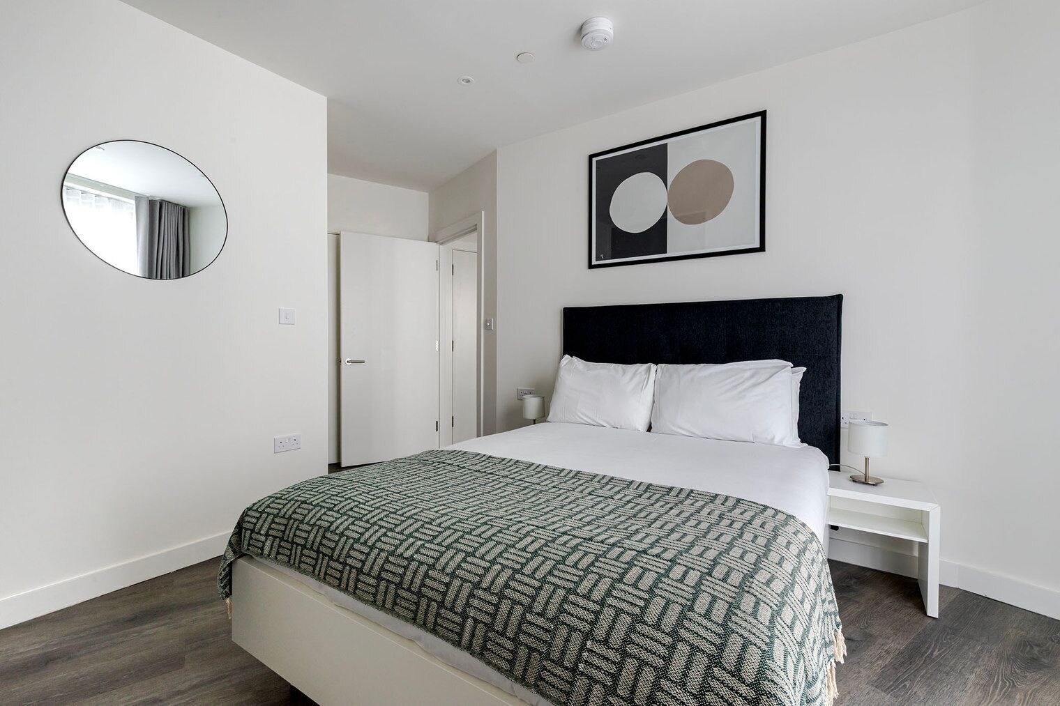 Een moderne hotelkamer met een groot bed, een ronde spiegel, een lijst met abstracte kunst en een witte nachtkastje.