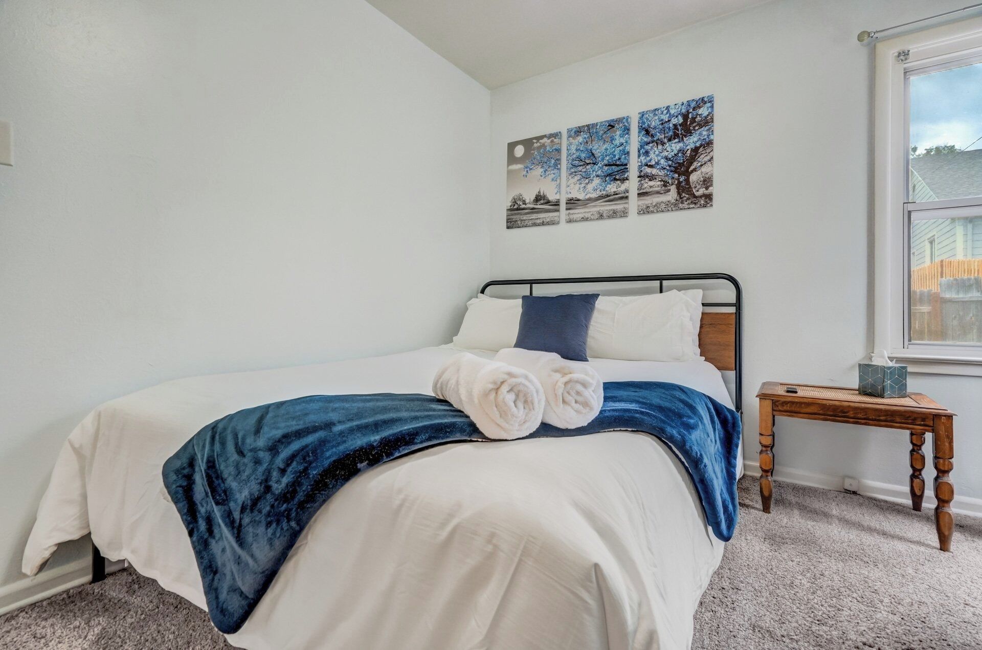 A neatly made bed with white and blue bedding, a wooden bedside table with a tissue box, and a window with a view of a fence.