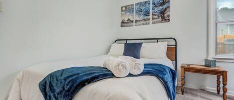 A neatly made bed with white and blue bedding, a wooden bedside table with a tissue box, and a window with a view of a fence.