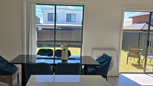 Un coin repas avec une table et des chaises en verre, un vase rempli de fleurs et une vue sur une cour arrière avec une clôture et une maison.