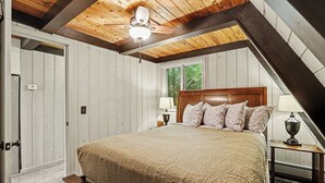 Un dormitorio con techo de madera, una cama con ropa de cama estampada, una mesita de noche con lámpara y una ventana con vistas a árboles.