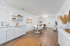 Une cuisine moderne avec des meubles blancs, un coin repas doté d’une table ronde et de chaises, et un salon avec un canapé et un téléviseur.