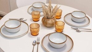 A round dining table with white plates and bowls, accompanied by matching cutlery and amber-colored glassware.