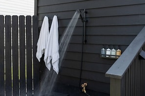 A showerhead with water flowing, a wooden fence, and a wall-mounted shelf with bottles.