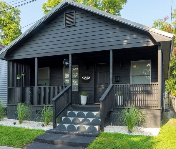 Une maison d’un seul étage, de couleur gris foncé, avec un porche à l’avant, un escalier menant à l’entrée et une enseigne sur laquelle on peut lire « casa ».