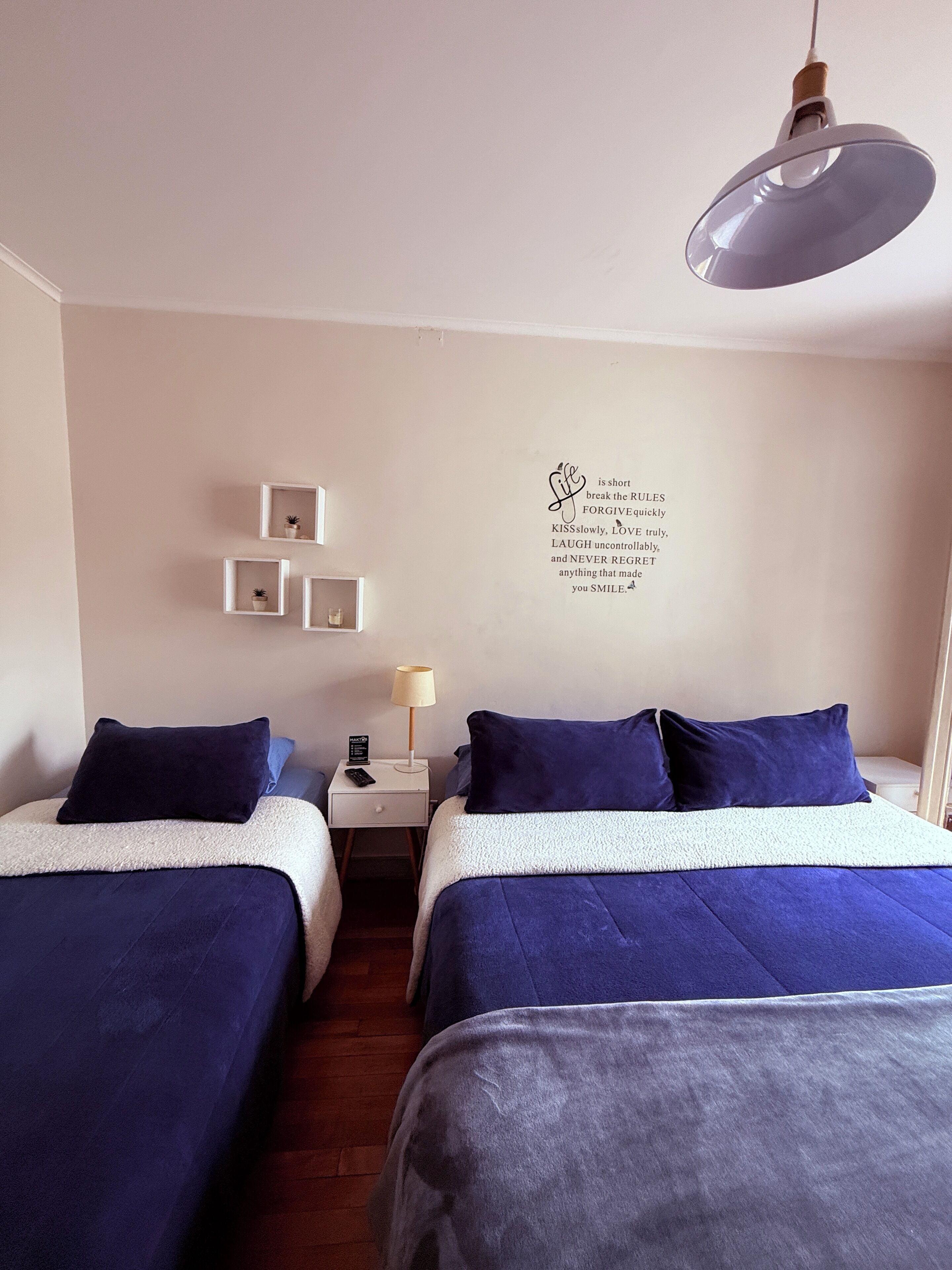Une chambre d’hôtel avec deux lits, une table de chevet avec une lampe, une étagère murale avec des plantes en pot et une citation inspirante sur le mur.