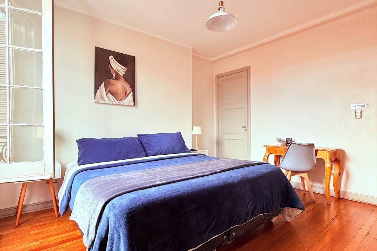A bedroom with a bed, a desk, a chair, a painting, and a window.