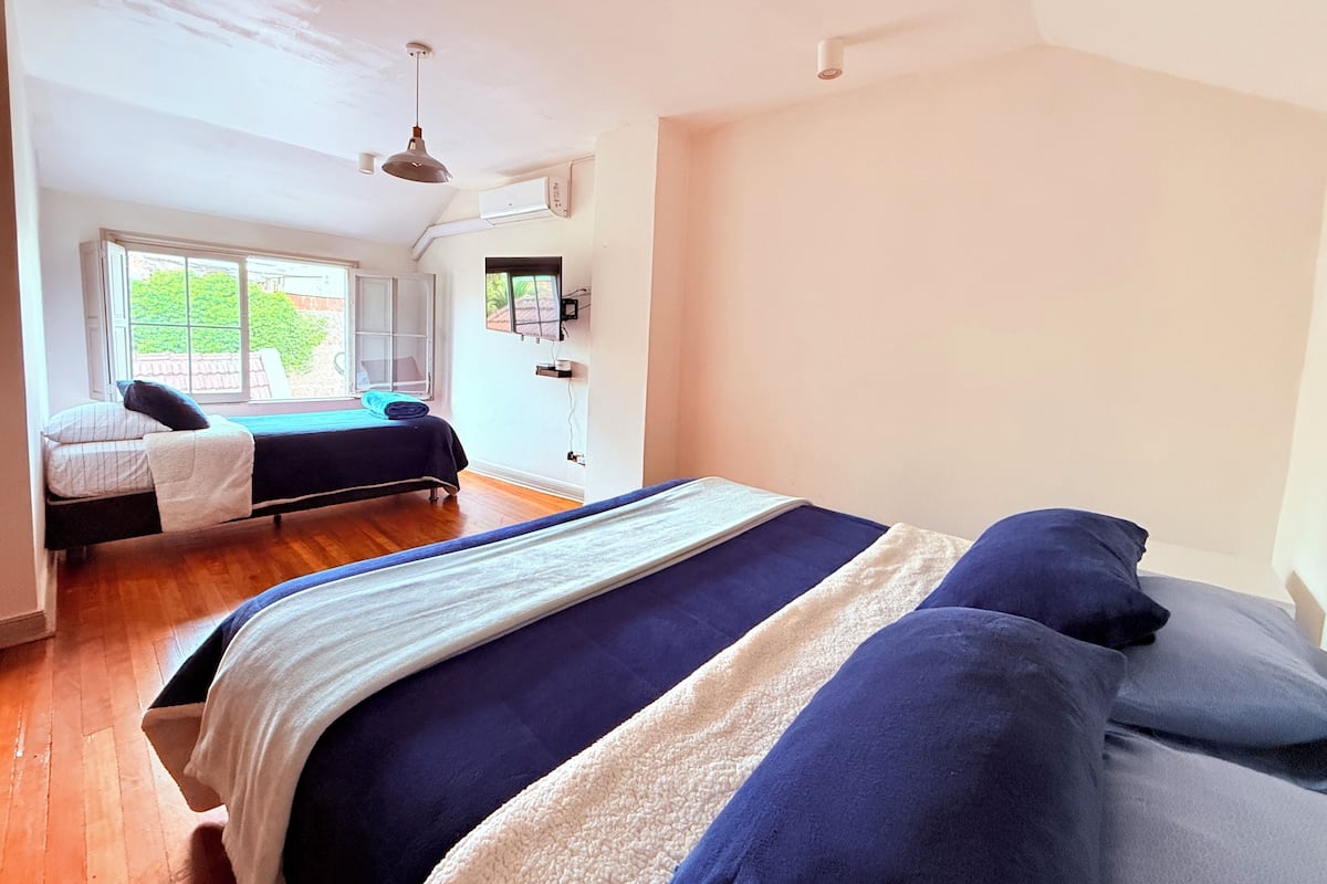 A bedroom with a large bed, a window with a view of greenery, a ceiling fan, and an air conditioning unit.