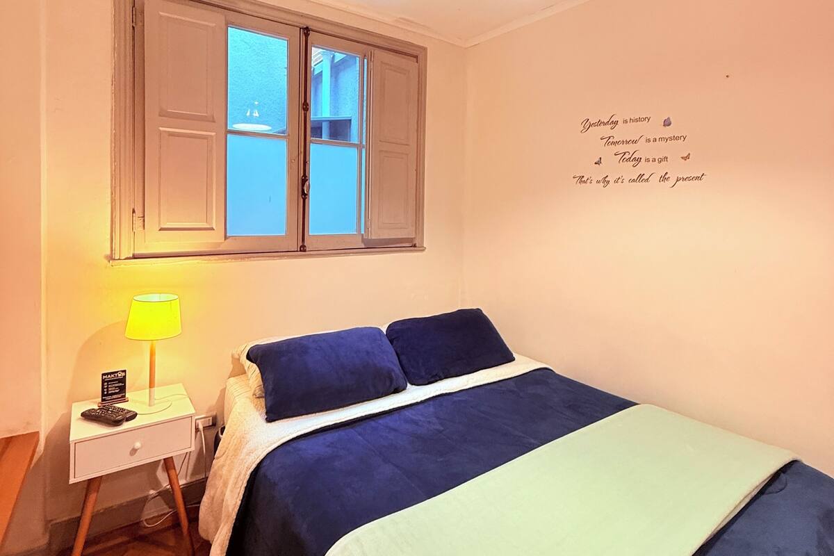 A bedroom with a bed, a nightstand, a lamp, a window, and a wall with a motivational quote.