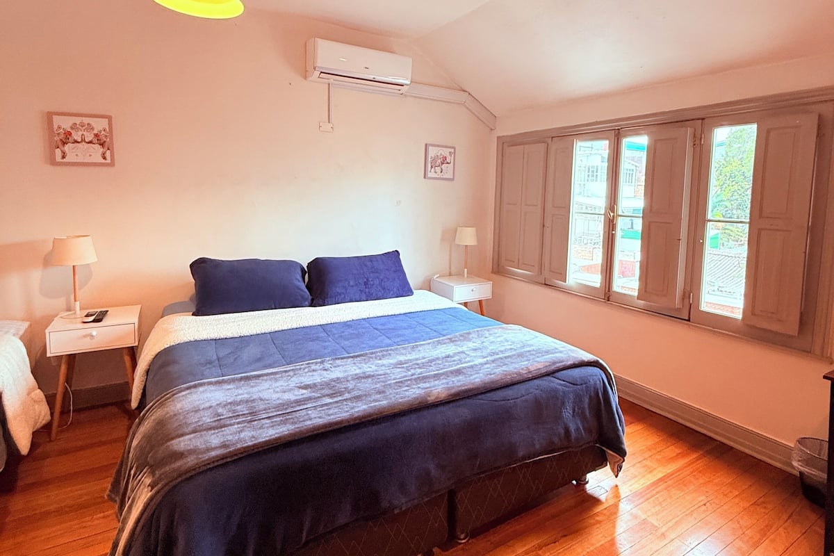 A bedroom with a large bed, two bedside tables, an air conditioning unit, and a window with shutters.