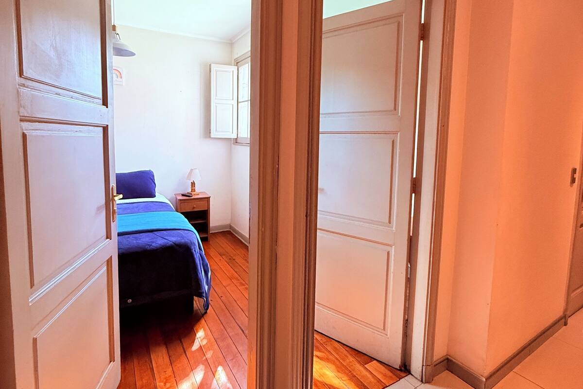 A hallway leading to a bedroom with a wooden floor, a bed with a blue blanket, and a nightstand with a lamp.
