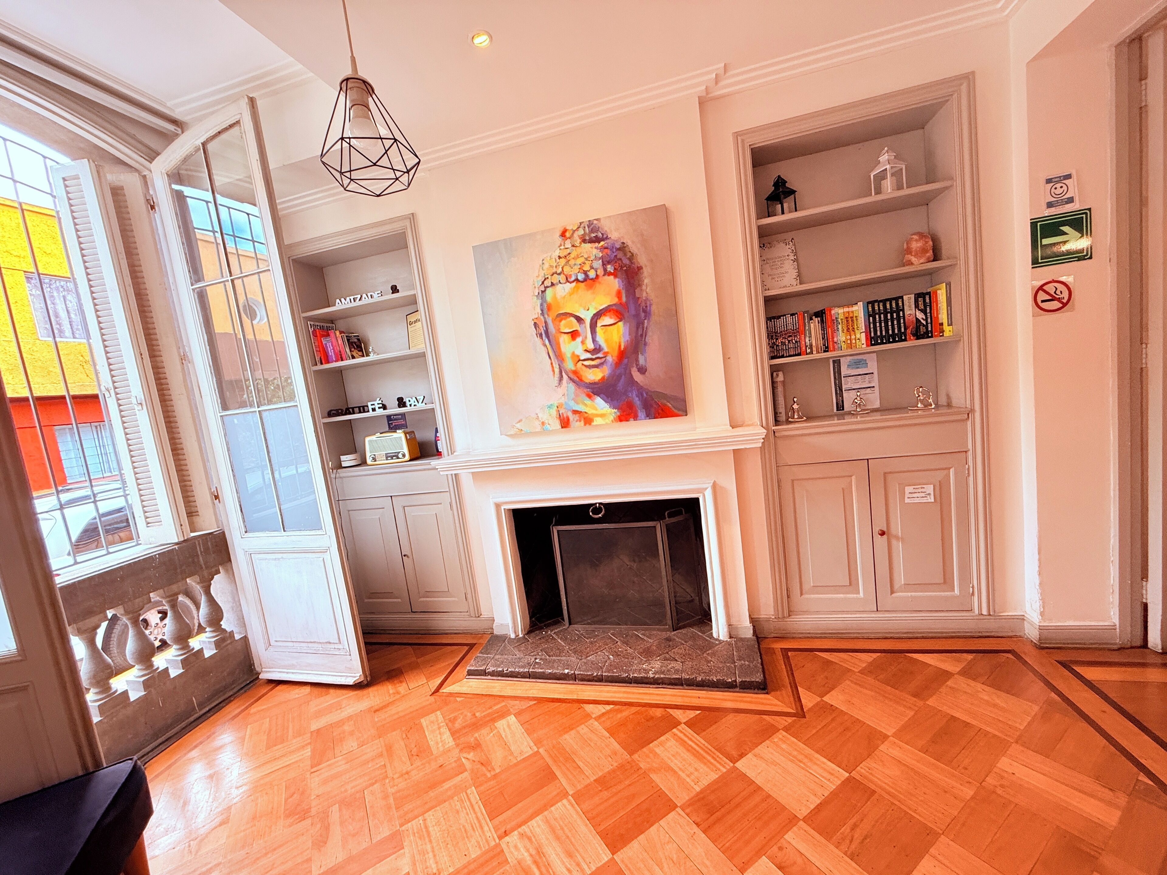 A room with a fireplace, a Buddha painting, bookshelves, and a window with shutters.