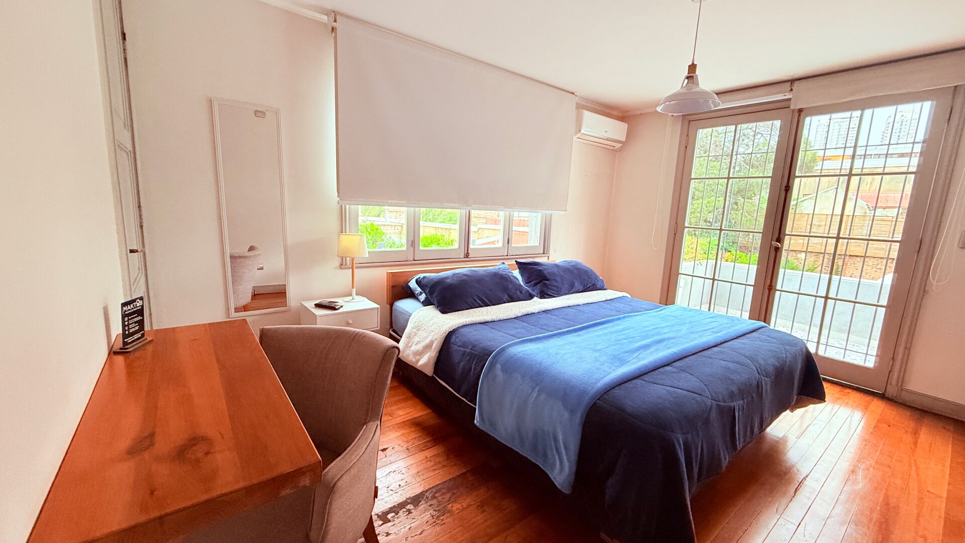 Ein Schlafzimmer mit Bett, Schreibtisch, Stuhl, Nachttisch, Lampe und einem Fenster mit Blick ins Grüne.