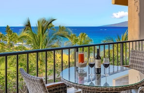 Un balcon avec une table en verre, des chaises en osier et une vue sur l’océan et la plage.