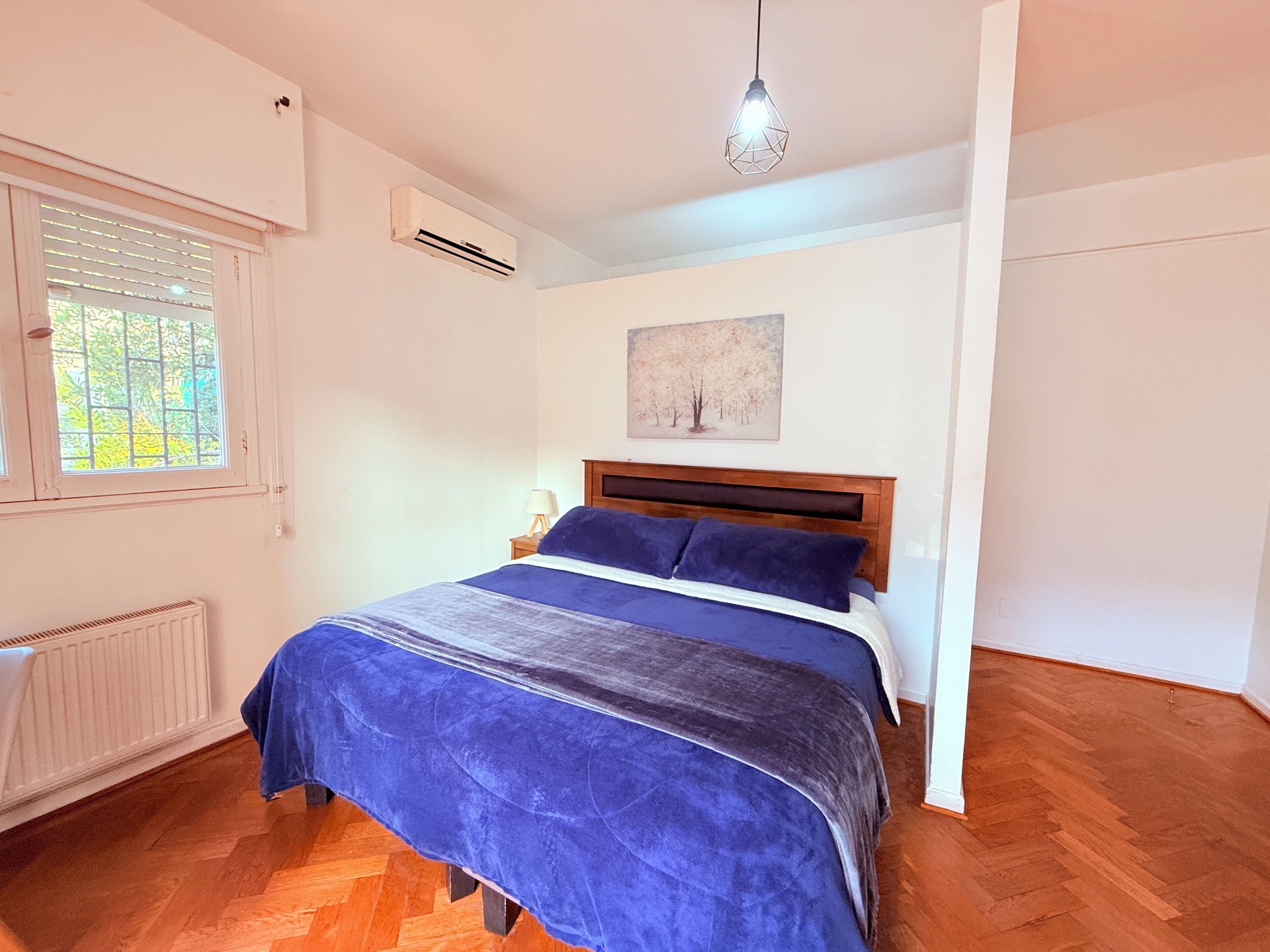 A bedroom with a bed, a radiator, a window, an air conditioner, and a hanging light fixture.