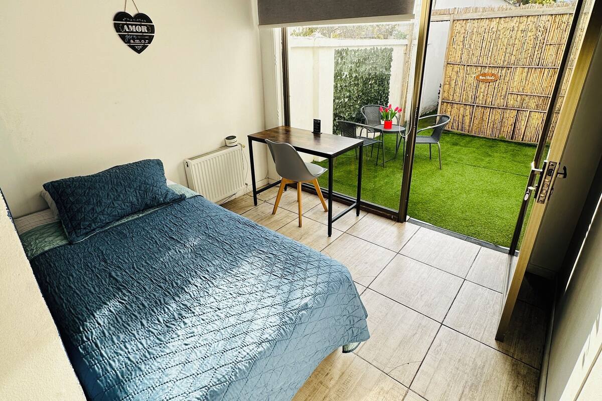 Ein Schlafzimmer mit Bett, Schreibtisch und Blick auf einen Garten durch eine Schiebetür.