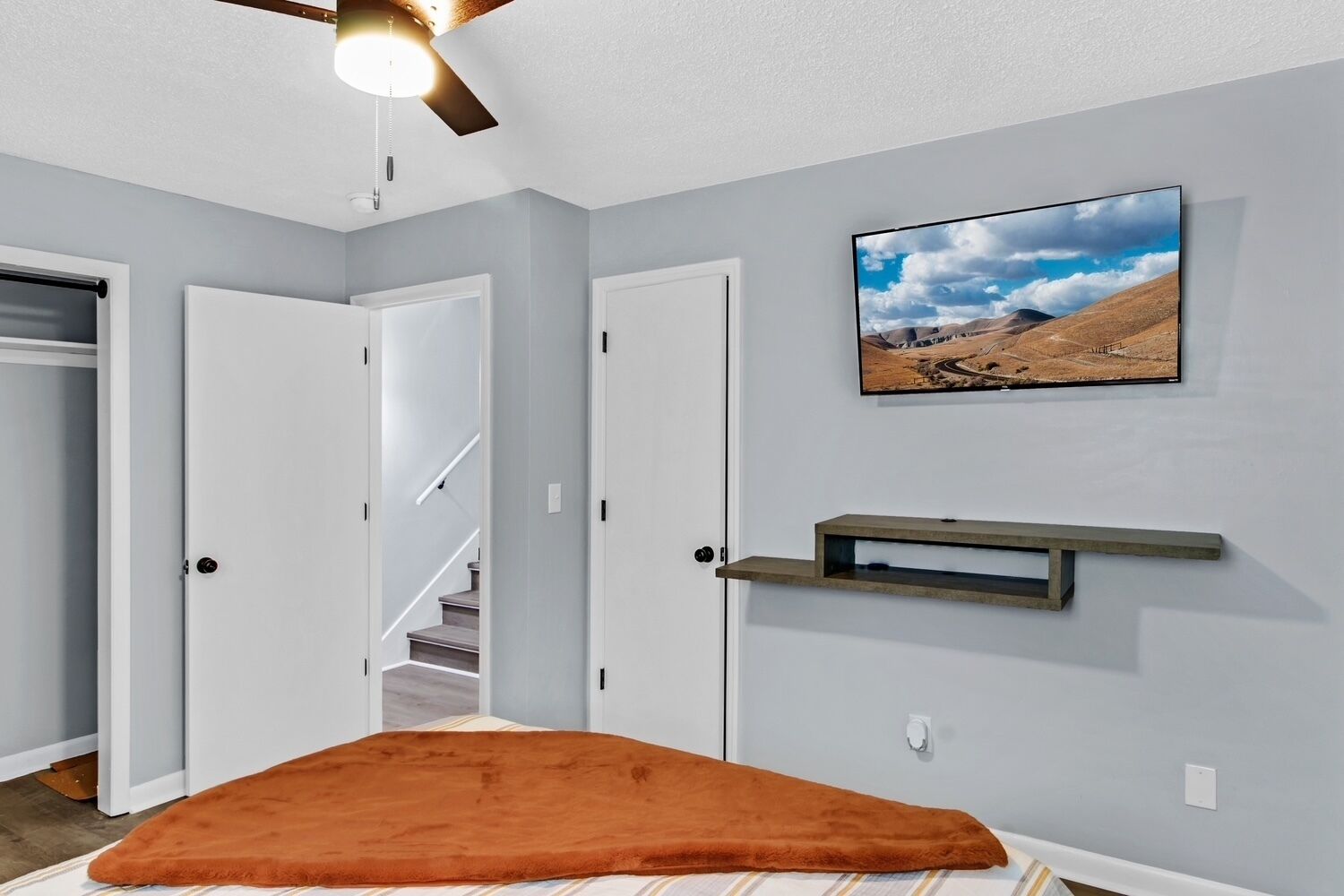 Une chambre à coucher avec un lit, un ventilateur de plafond, un téléviseur fixé au mur et un couloir menant à une autre pièce.