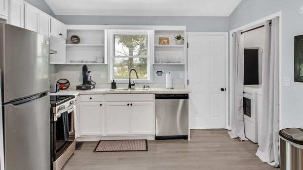 A modern kitchen with white cabinets, a stainless steel refrigerator, a dishwasher, and a window with blinds.