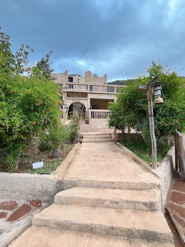A building with a stone staircase leading to a gated entrance, surrounded by lush greenery and a clear sky. - Gite jawaher aguerd (Aqesri)