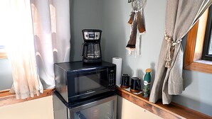 Een kleine keuken met een magnetron en koffiezetapparaat op een aanrecht, een koelkast, en een raam met gordijnen.