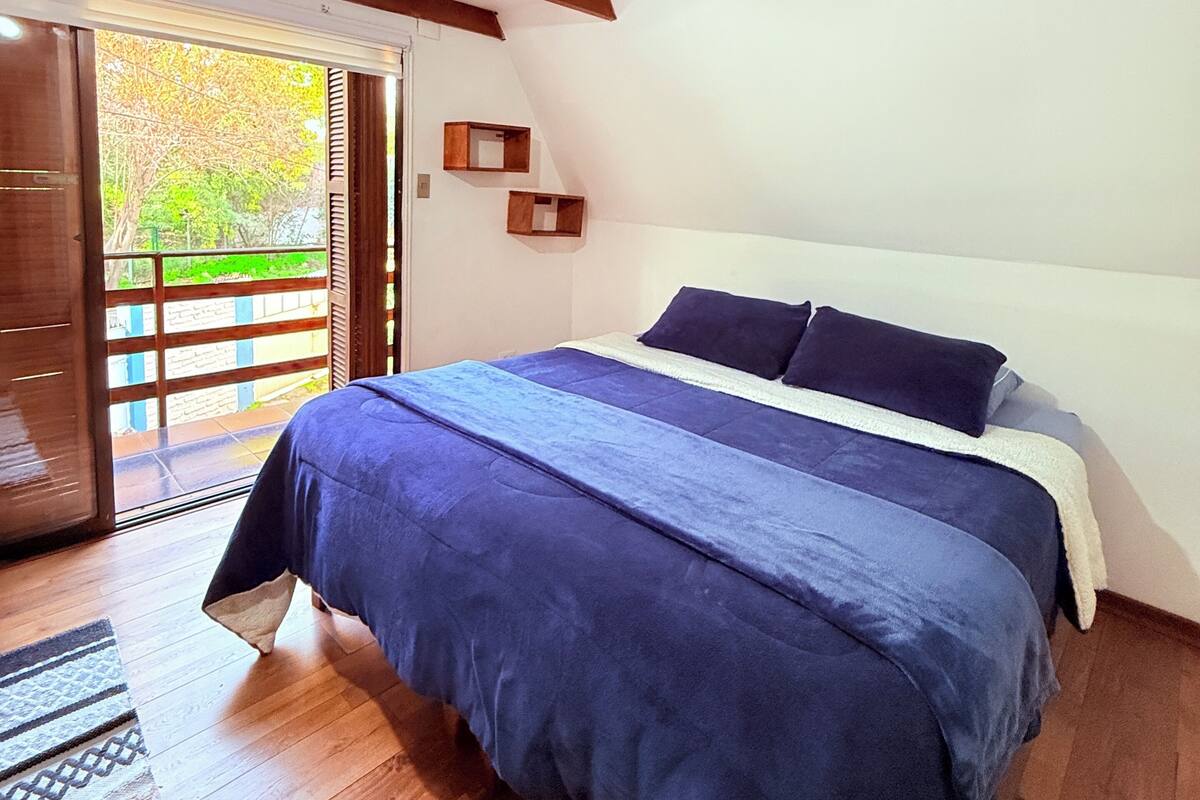 Une chambre avec un grand lit, des poutres en bois, une fenêtre donnant sur un paysage verdoyant et une porte menant à l’ext érieur.
