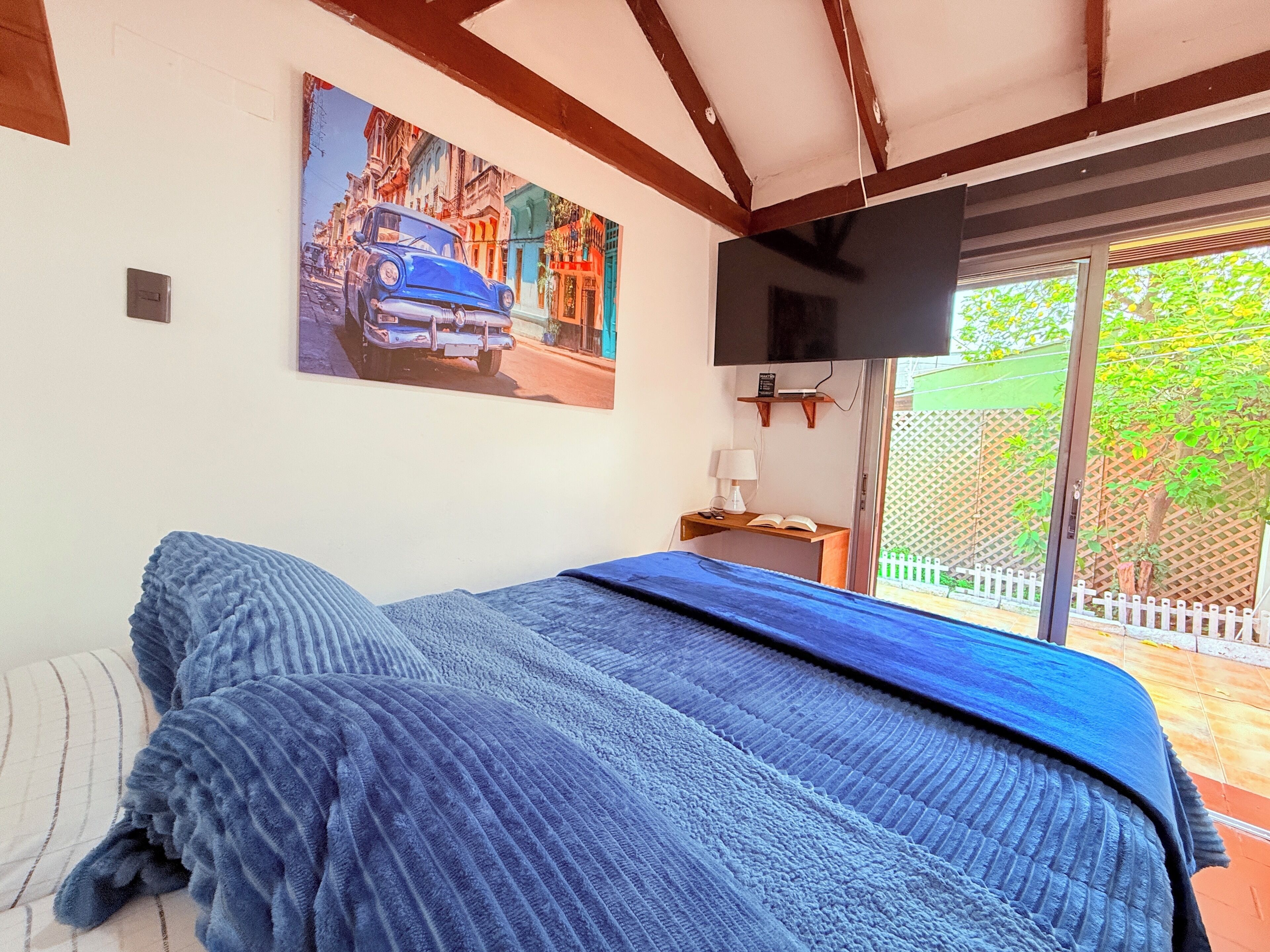 A bedroom with a bed, a painting of a car on the wall, a TV mounted above the bed, and a window with a view of greenery outside.
