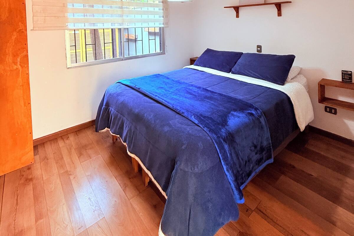 Une chambre à coucher avec un lit, un plancher en bois, une fenêtre avec des stores, un luminaire suspendu et une petite étagère avec un panneau.