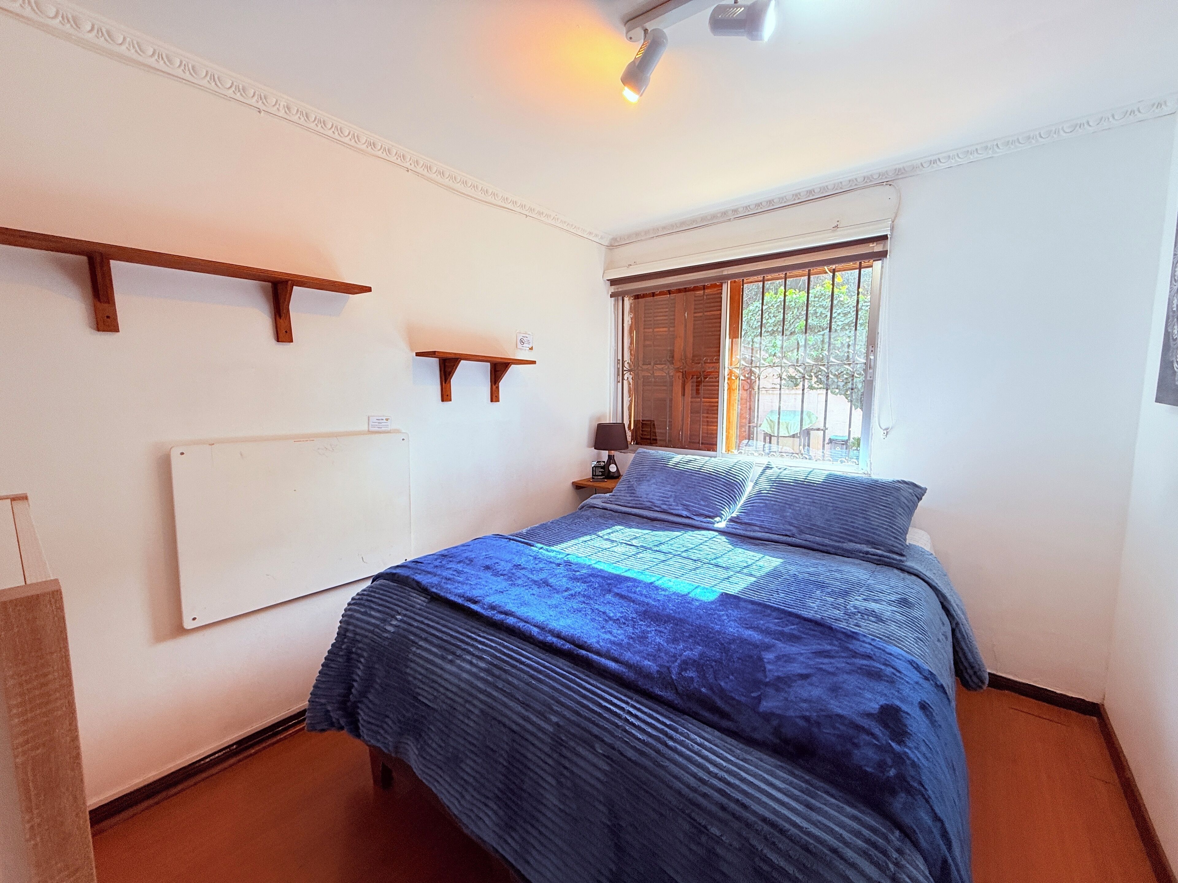 A bedroom with a bed, a nightstand, a window with blinds, and a wall-mounted shelf.