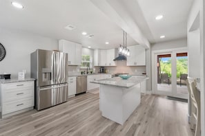 A modern kitchen with a stainless steel refrigerator, white cabinetry, a granite countertop island, and a view to the outdoors.