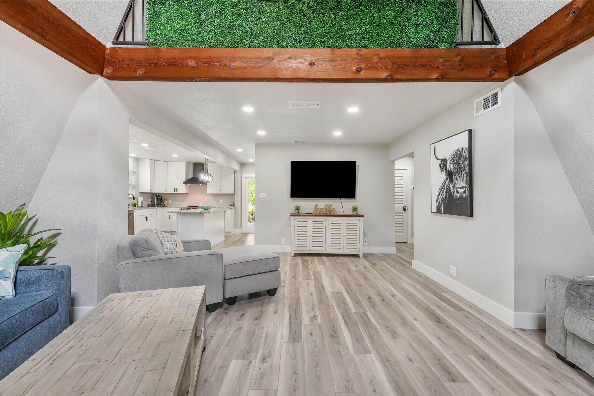 Un salon moderne avec un plancher en bois, des canapés gris, une table basse, un téléviseur et une cuisine en arrière-plan.