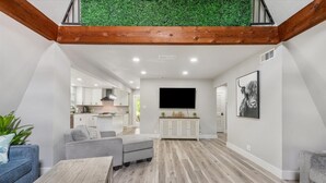 Un salon moderne avec un plancher en bois, des canapés gris, une table basse, un téléviseur et une cuisine en arrière-plan.