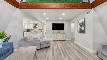 Un salon moderne avec un plancher en bois, des canapés gris, une table basse, un téléviseur et une cuisine en arrière-plan.