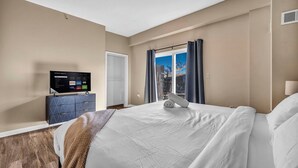 A hotel room with a bed, a TV on a dresser, a window with curtains, and a view of the outside.