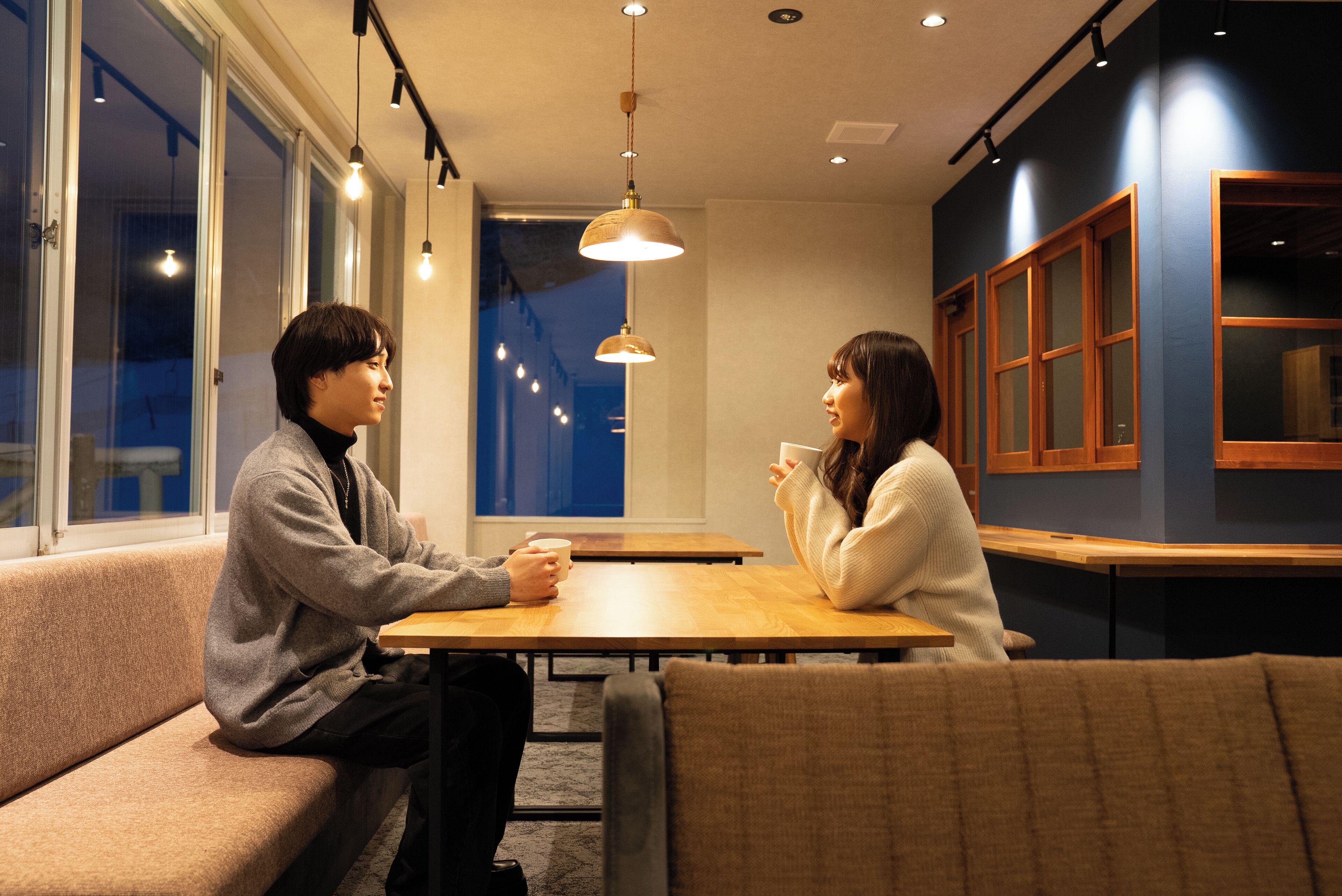 A modern interior with a wooden table and bench seating, large windows, and pendant lighting.