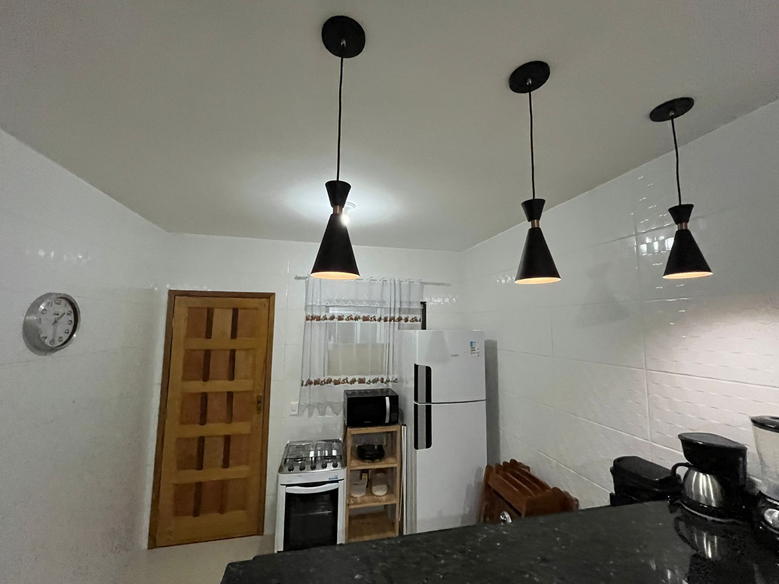 Una cocina con paredes de azulejos blancos, una puerta de madera, una estufa, un refrigerador y tres lámparas colgantes.