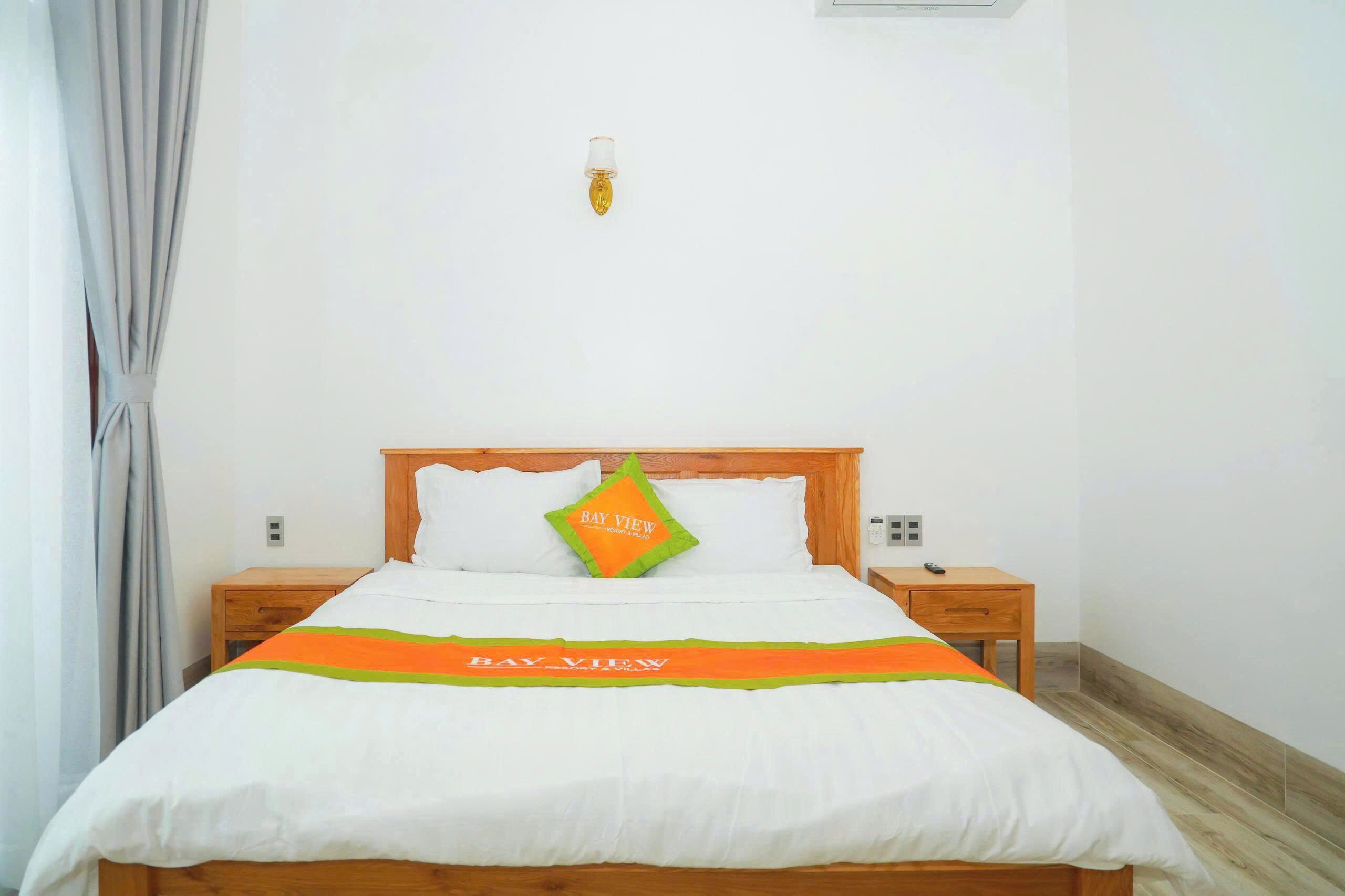 Ein ordentlich bezogenes Bett mit einer weißen und orangefarbenen Tagesdecke, ein Kopfkissen mit dem Aufdruck "Bay View" und ein Nachttisch mit einer Lampe.