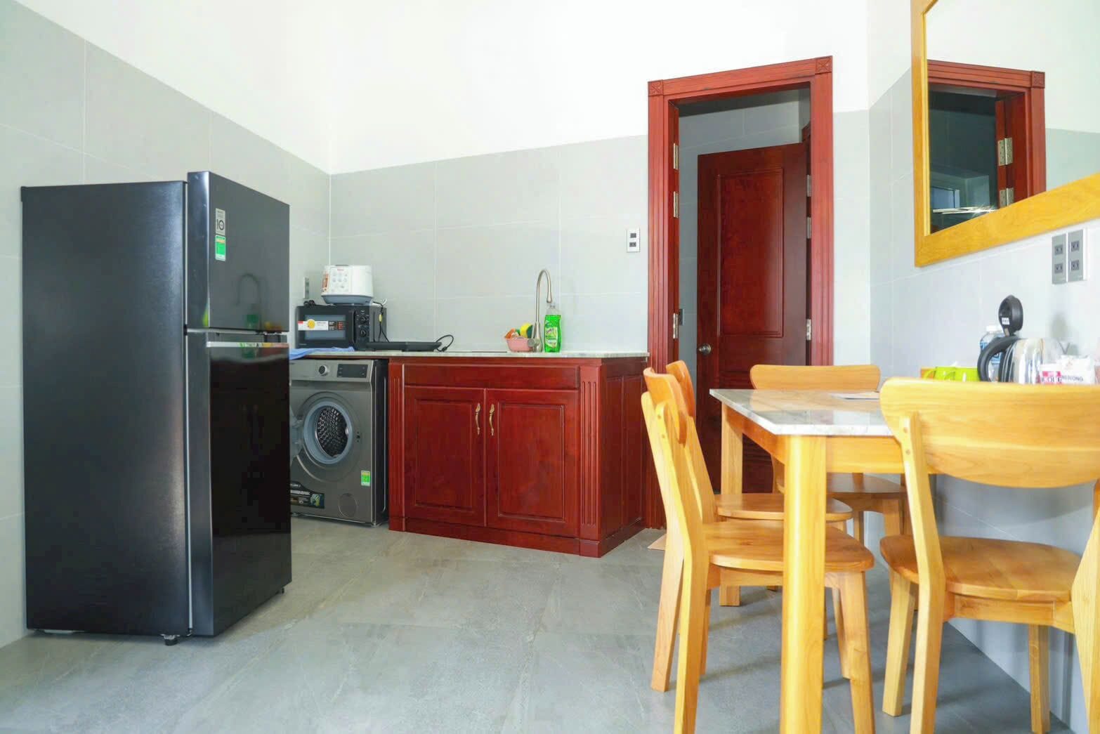 A compact kitchen with a refrigerator, microwave, washing machine, wooden cabinets, and a dining table with chairs.