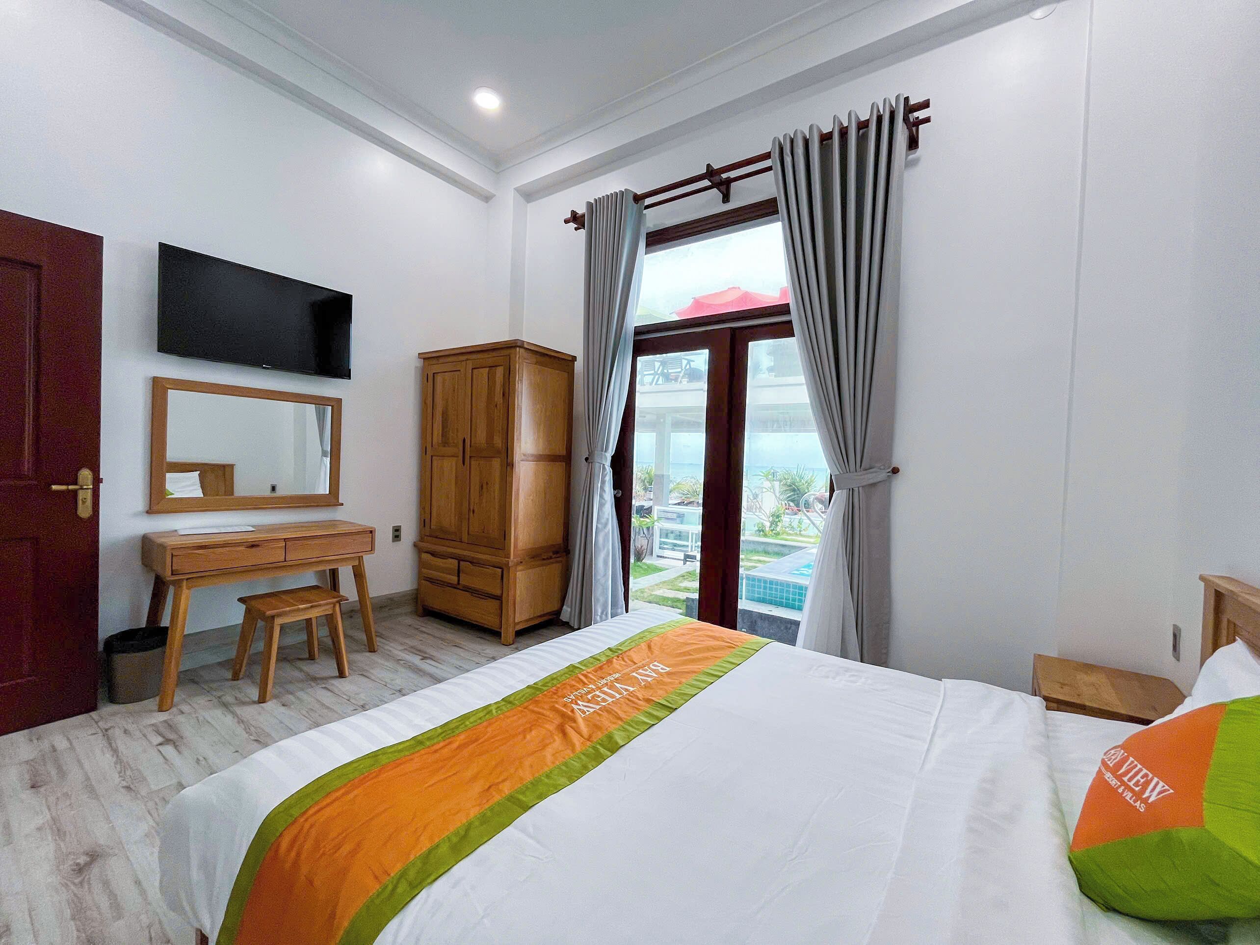 A hotel room with a bed, a wooden desk, a mirror, a wardrobe, a TV, and a window with curtains.
