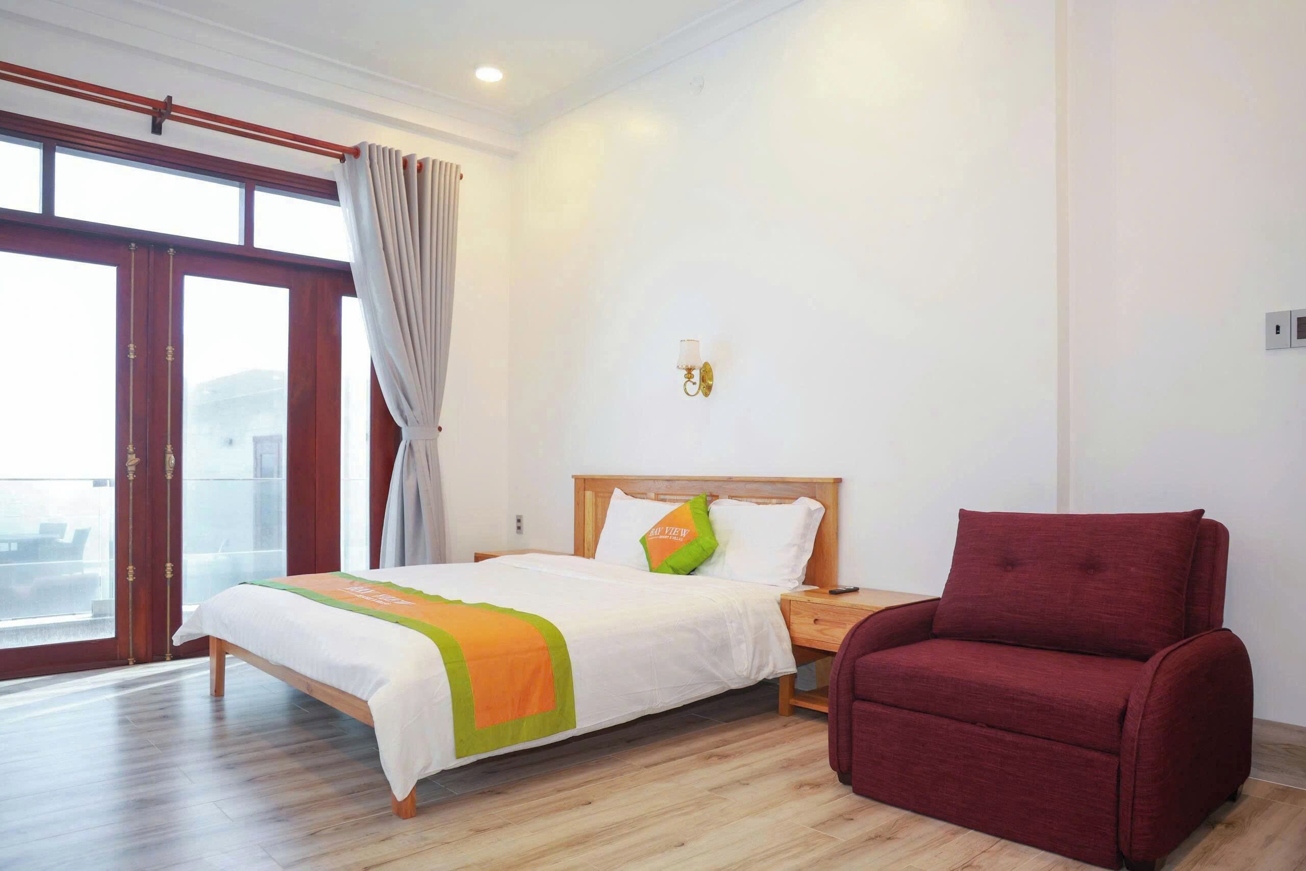A hotel room with a bed, a red armchair, a nightstand, a wall-mounted lamp, and a large window with curtains.