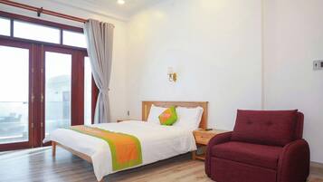 A hotel room with a bed, a red armchair, a nightstand, a wall-mounted lamp, and a large window with curtains.
