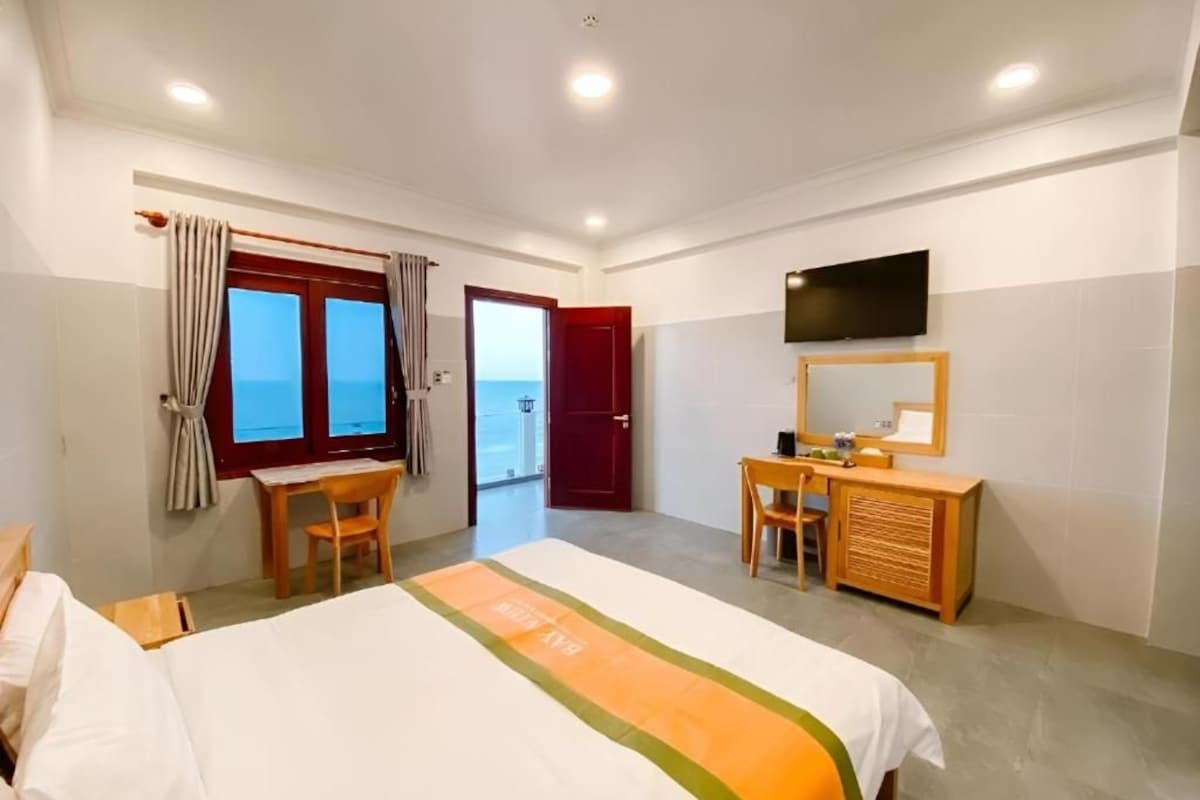 A hotel room with a bed, a desk, a chair, a TV, and a view of the sea through the window.
