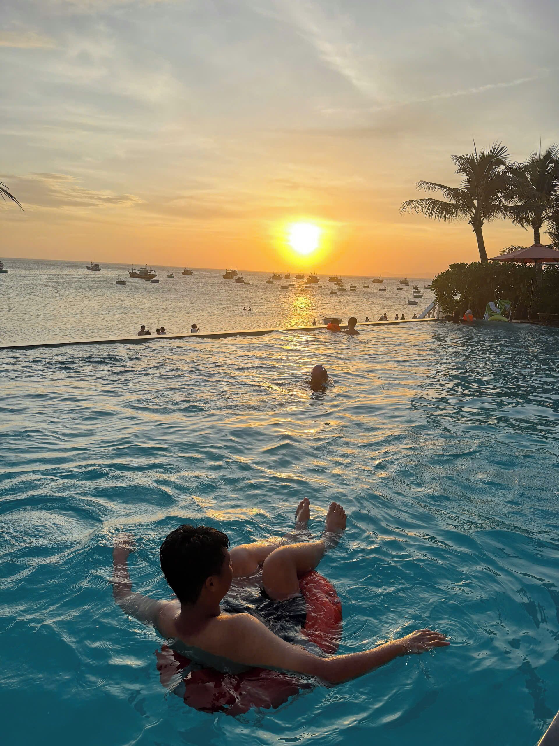 A pool with people swimming and relaxing, a sunset in the background, and boats on the water.