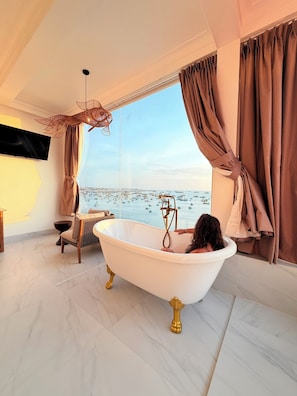 A modern bathroom with a freestanding bathtub, a large window with a view of the sea, and a decorative ceiling light. - BayView Resort & Villas (Mui Ne)