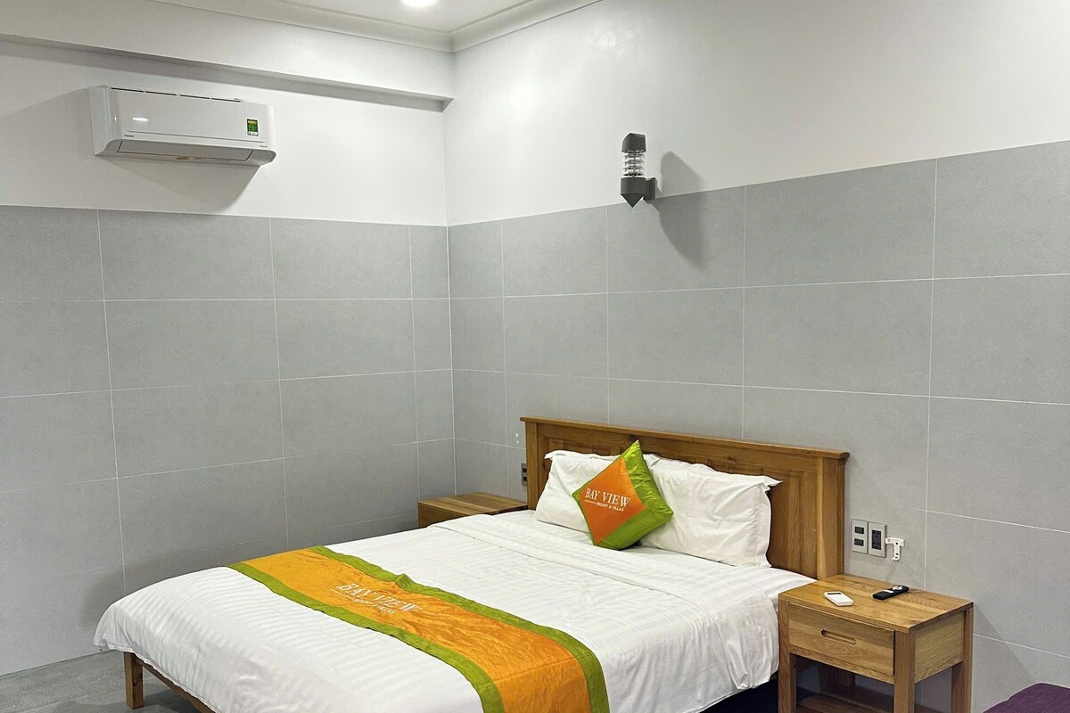 A hotel room with a bed, a nightstand, a purple chair, a wall-mounted air conditioner, and a wall-mounted light fixture.