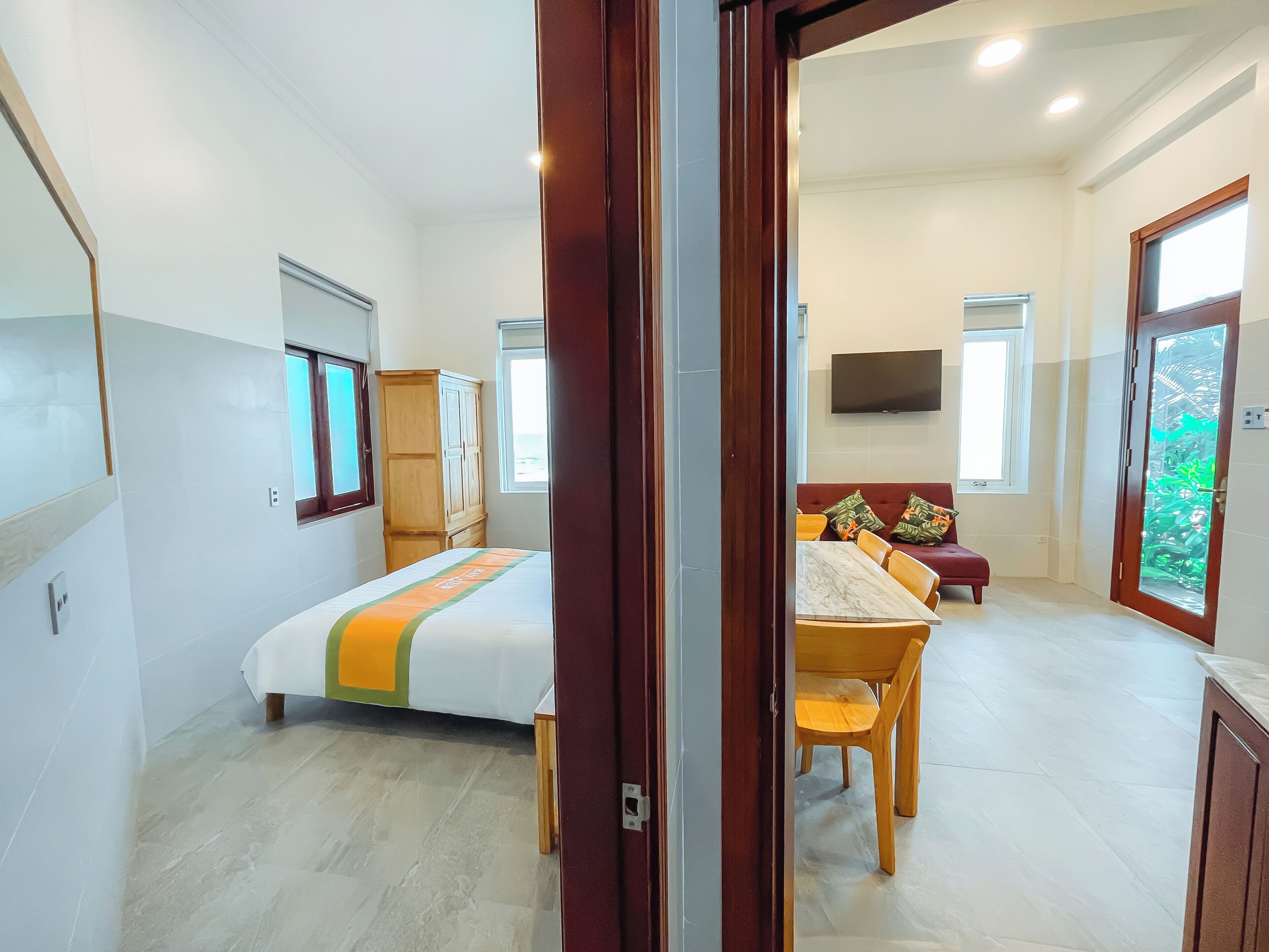 A hotel room with a bed, a wooden wardrobe, a dining table with chairs, a television, and a large window.