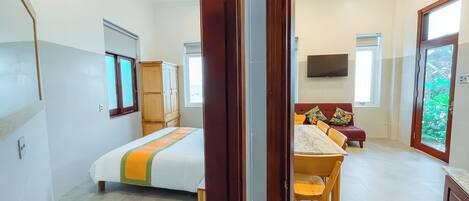 A hotel room with a bed, a wooden wardrobe, a dining table with chairs, a television, and a large window.