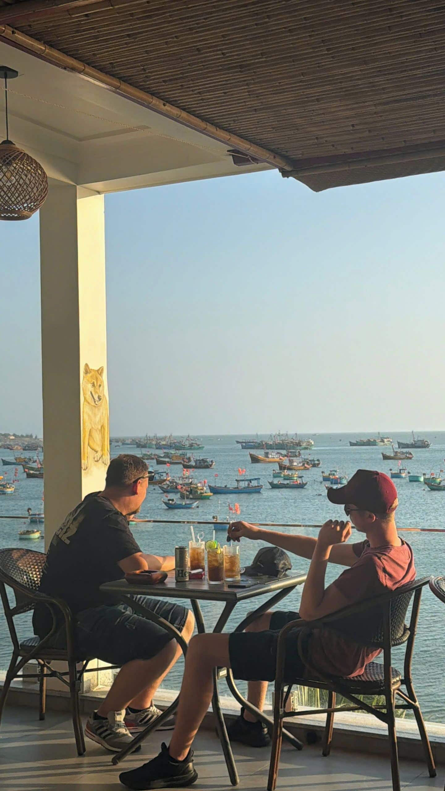 Duas pessoas sentadas a uma mesa com bebidas, com vista para um porto cheio de barcos.