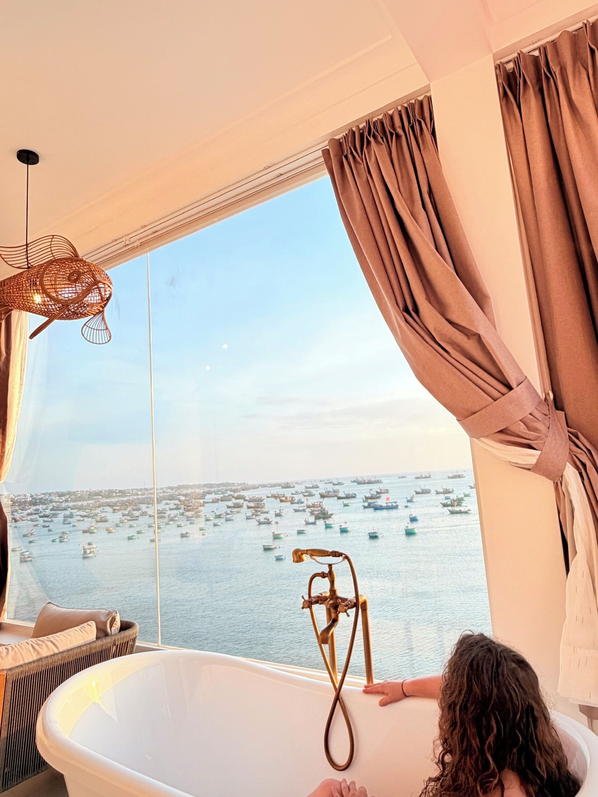 A bathroom with a bathtub, a window with curtains, and a view of boats on the water.