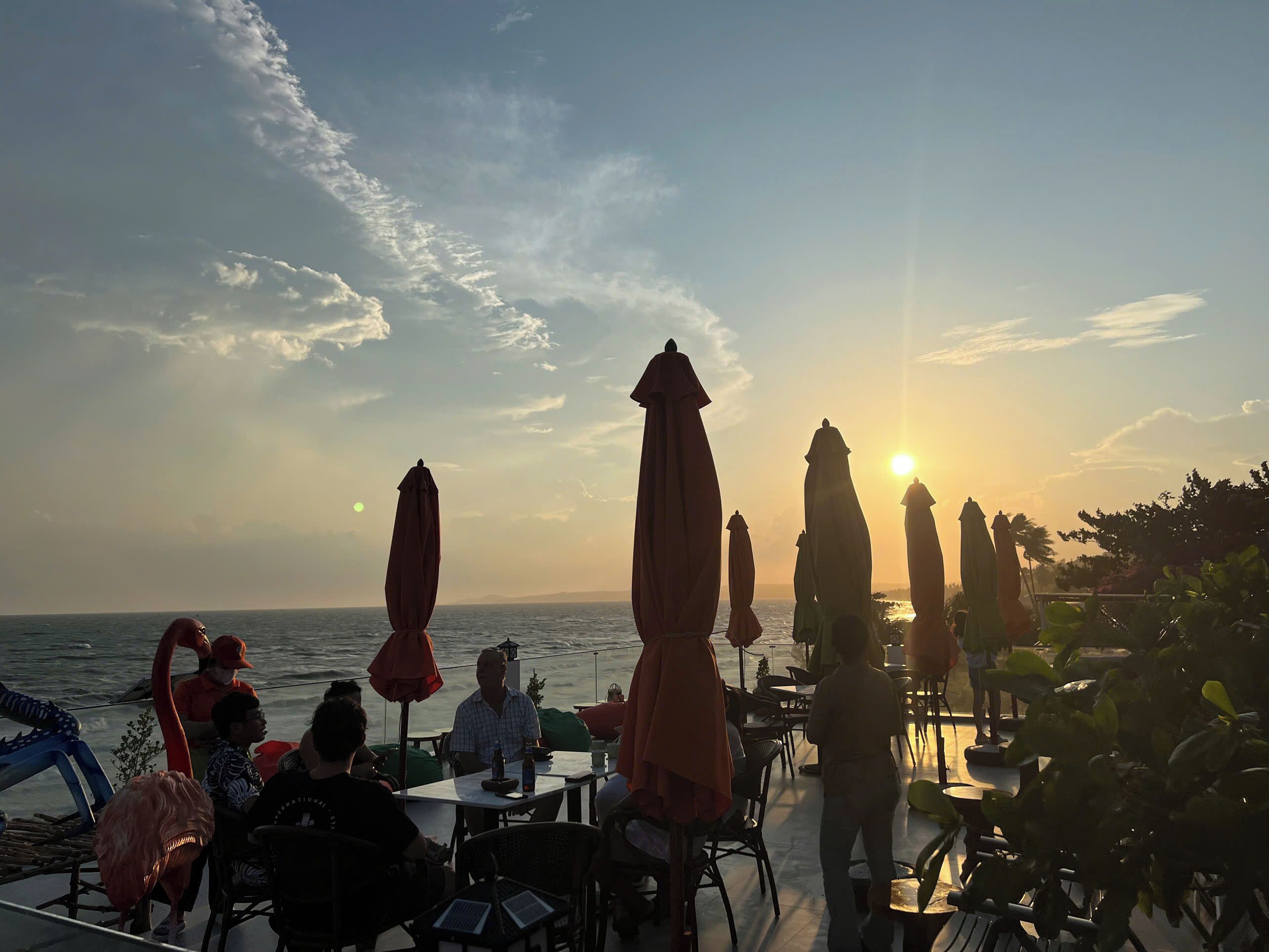 A seaside restaurant with outdoor seating, overlooking the ocean, featuring umbrellas and tables.